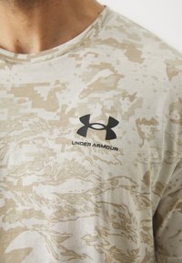 Beige camouflage T-shirt med korte ærmer, lavet af bomuld. Har et sort Under Armour-logo på forsiden. Blød tekstur, design med rund hals.