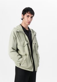 Light green jacket with a hood, front zip closure, two large chest pockets, and elastic cuffs. Features a windbreaker design and textured fabric.