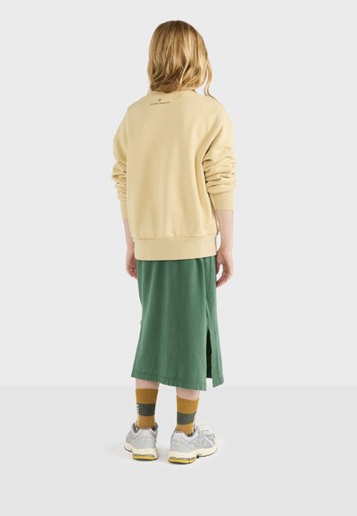 Kremowy sweatshirt o luźnym kroju, połączony z długą zieloną spódnicą z bocznym rozcięciem. Srebrne sneakersy i musztardowe skarpetki dopełniają stylizację.
