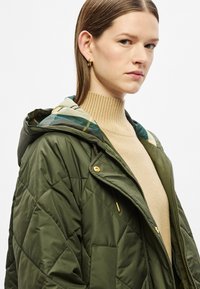 Olivově zelená prošívaná bunda s kapucí, která má vzorovanou podšívku, zlaté kovové detaily, zapínání na zip a přední kapsy.