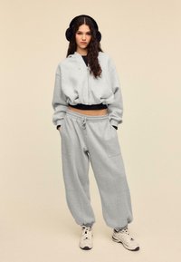 Grå cropped hættetrøje med lynlås foran, kombineret med matchende løse sweatpants. Blød tekstur, elastisk taljebånd og sidelommer. Hvide sneakers.