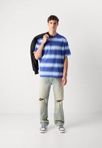 Camiseta a rayas azul con mangas cortas, combinada con jeans claros rasgados. El modelo sostiene una chaqueta negra, vistiendo zapatillas con acentos azules.