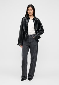 Γυναίκα που στέκεται φορώντας ένα μαύρο δερμάτινο μπουφάν πάνω από λευκό τοπ, σκούρα τζιν με φαρδιά πόδια και μαύρα μυτερά παπούτσια, μπροστά σε απλό φόντο.