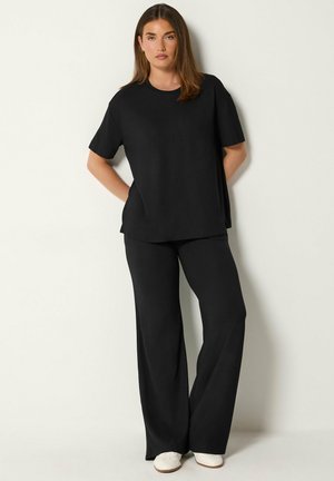 Femme debout contre un mur clair, portant un haut noir ample à manches courtes, un pantalon large noir et des chaussures blanches, mains dans le dos.