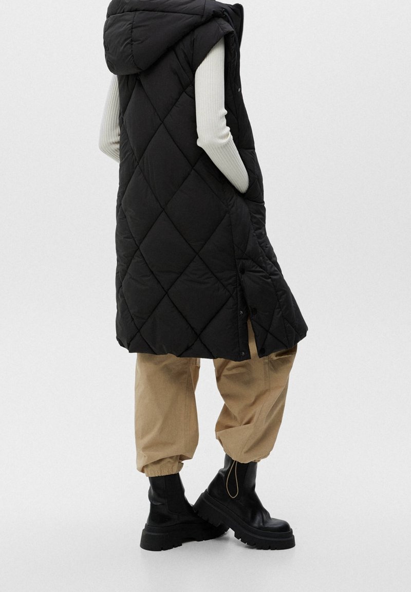 Personne portant une longue veste matelassée noire avec une capuche, un pantalon beige à revers, des bottes noires épaisses et un haut blanc côtelé à manches longues.