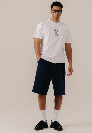 Άνδρας που φοράει λευκό t-shirt με γραφικά, σκούρα σορτς μέχρι το γόνατο, λευκές κάλτσες, μαύρα παπούτσια και σκούρα γυαλιά ηλίου, στέκεται με το ένα χέρι στην τσέπη.