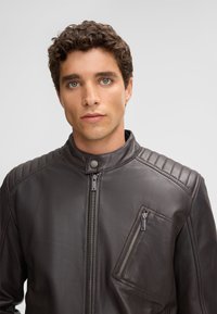 Braune Leder-Motorradjacke mit gesteppten Schulterakzenten, hohem Kragen und asymmetrischem Reißverschlussfach auf der Brust. Glatte Textur.