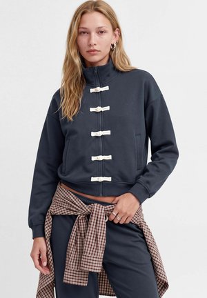 Femme aux longs cheveux blonds porte une veste bleu marine avec des détails blancs à boutons à bascule et une chemise marron à carreaux nouée autour de la taille.
