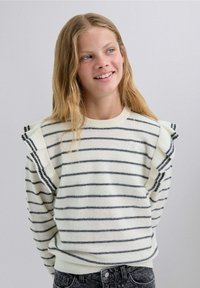 Randig stickad tröja i cream och svart, med volangdetaljer på axlarna och rund halsringning, tillsammans med mörka, strukturerade jeans.
