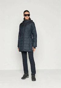 Kvinde iført en lang sort quilted frakke, sorte bukser, sorte vandrestøvler og store sorte solbriller, stående foran en lys ensfarvet baggrund.