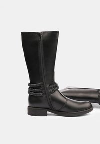 Botas negras hasta la rodilla hechas de material sintético suave, con cierre de cremallera y una correa trenzada negra decorativa alrededor del tobillo.