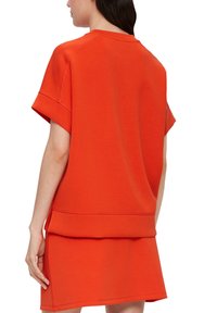 Vrouw in een losse, feloranje top met korte mouwen en bijpassende rok, gezien vanaf de achterkant met ontspannen armen langs haar zij.