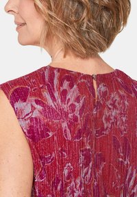 Blumenkleid in Rottönen mit strukturiertem Stoff. Verfügt über einen Ausschnitt im Rücken und ist ärmellos.