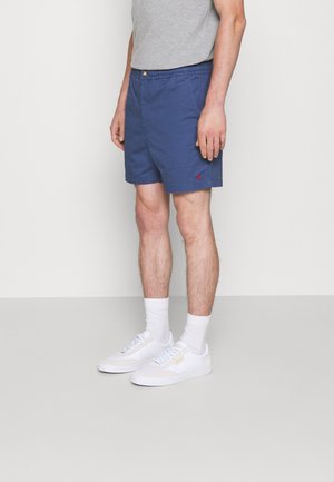 Man die marineblauwe shorts, een grijs T-shirt, witte sokken en witte sneakers draagt, staande tegen een effen achtergrond.