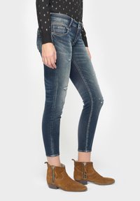 Jeans ajustados de denim, con un degradado de azul claro a oscuro, con costuras visibles y desgaste en las rodillas. Acompañados de botas de tobillo marrones con cremalleras laterales.