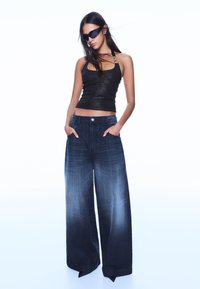 Černý lesklý top bez rukávů spojený s tmavými džínami s širokými nohavicemi. Džíny mají přední kapsy a zapínání na knoflík.