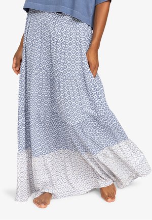 Femme portant une jupe longue fluide à motifs floraux bleus et blancs avec une taille élastique, debout pieds nus devant un fond blanc.