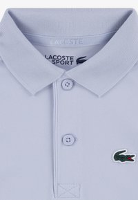 Ανοιχτό γαλάζιο πόλο πουκάμισο από υφασμάτινο υλικό με υφή, με γιακά που διπλώνει, δύο κουμπιά και πράσινο λογότυπο κροκόδειλου στο στήθος.