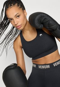 Melns sporta krūšturis ar apaļu izgriezumu un nelielu logotipu. Kombinēts ar melnām legingām ar augstu vidukli, uz kurām vidukļa joslā redzams "VENUM" zīmols.