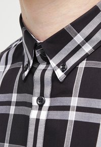 Närbild av en skjorta med krage som har ett svartvitt rutmönster, med synliga knappar och en del av en individs hals.