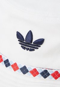 White bucket hat featuring a navy embroidered three-stripe logo and a multicolored argyle patterned band in red, blue, and black.