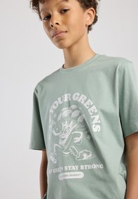 Mint green cotton t-shirt featuring a playful white graphic of broccoli with arms and legs, text reads "EAT YOUR GREENS STAY STRONG."