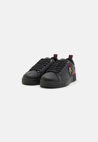 Black sneakers made of synthetic leather with flat laces, rounded toe, textured side panels, and a colorful embroidered patch on the outer side.