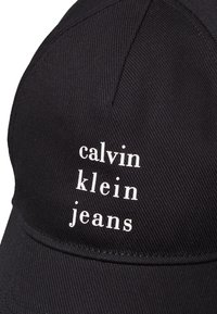 Juoda beisbolo kepurė su išlenktu kraštu, priekyje esantis baltas užrašas "calvin klein jeans", lygus audinys su įstrižine šonkauline tekstūra.