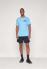 Mees seisab seljas helesinine Nike shirt, mustad sportlikud lühikesed püksid, mustad sokid ja mustad spordijalatsid, planeedi valgusel taustal.