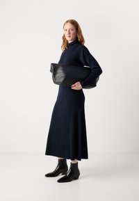 Vestido de punto acanalado en azul marino con cuello alto, combinado con un gran bolso de mano negro de cuero y botines negros con tacón. Texturas suaves y diseño moderno.