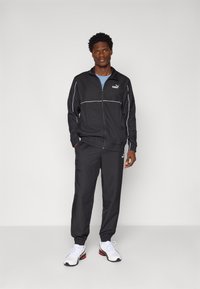 Black athletic jacket with white accents and logo, paired with matching pants. Model wearing a light blue T-shirt and white sneakers.
