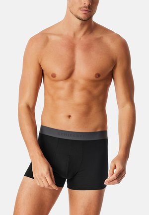 Schwarze Boxershorts mit einem grauen Bund, der geprägtem Text zeigt. Der Stoff wirkt glatt und eng anliegend und betont einen muskulösen Körperbau.