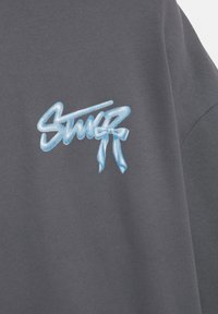 Temno siva tkanina z modrim stiliziranim napisom v grafitnem slogu "Stüssy" natisnjenim na površini.