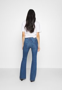 Farkut, joissa on levenevät lahkeet, sininen denim, keskikorkea vyötärö, takataskut, joissa on kaareva tikkaus, ja sileä pinta. Yhdistettynä yksinkertaiseen valkoiseen t-paitaan.