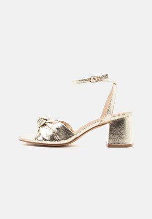 Sandale métallisée dorée à bout ouvert avec bride avant nouée, bride de cheville à boucle et talon bloc sur fond neutre.