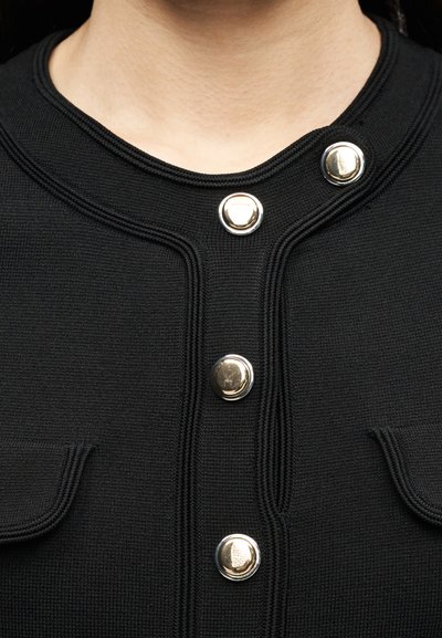 Κοντινό πλάνο μαύρου πλεκτού ενδύματος με τέσσερα ασημένια, λαμπερά κουμπιά και δύο μικρές τσέπες με καπάκι στο μπροστινό μέρος.