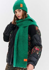 Zielona dzianinowa czapka z pomarańczową metką, pasujący zielony szalik, puchowa czarna kurtka w kwiatowe wzory i spodnie w panterkę.