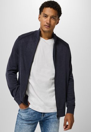 Marineblaue Zip-Jacke aus weichem Material mit gerippten Bündchen und Kragen, getragen über einem weißen T-Shirt, kombiniert mit hellblauen Jeans.
