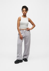 Γυναίκα που στέκεται με το χέρι στη μέση, φορώντας ανοιχτό γκρι αμάνικο crop top, άνετο γκρι φαρδύ παντελόνι φόρμας και μαύρα chunky sneakers σε λευκό φόντο.