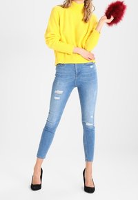 Yellow knit sweater with a round neck, paired with light blue skinny jeans featuring distressed details. Model holds a red fluffy accessory.