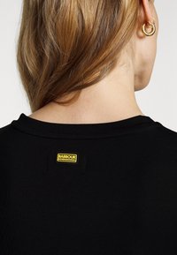 Černé tričko s kulatým výstřihem, na zadní straně zdobené žlutou nášivkou "Barbour International", hladká textilie a přiléhavý střih.