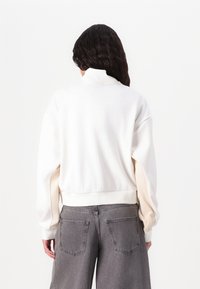 Hvid sweatshirt med høj krave og ribbet kant, der har beige detaljer på ærmerne; parret med grå brede bukser.