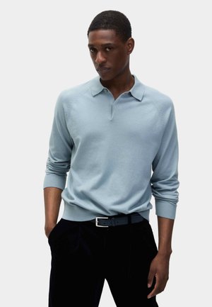 Lyseblå strikket pullover med krave og ribbede manchetter, båret med mørke bukser og et sort bælte. Glat tekstur, tætsiddende design.