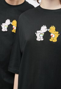 Μαύρο βαμβακερό μπλουζάκι με κεντημένα καρτουνίστικα αρκουδάκια—ένα λευκό με ουράνιο τόξο και ένα κ�ίτρινο με καρδιά—στο κέντρο του στήθους.