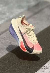 Tenisice s svijetlosmešanim i koraljnim gornjim dijelom, crnim Swoosh logom, teksturiranim pletenim materijalom, ljubičastom potplatom i jarkocrvenim akcentom na peti.