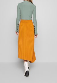 Haut vert à manches longues avec des accents de boutons, associé à une jupe midi plissée orange. Des bottes à talons blanches complètent la tenue, le tout sur fond gris.