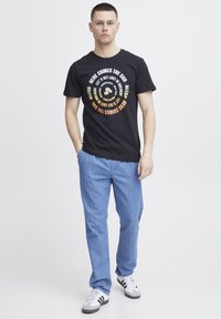 Svart bomull t-skjorte med sirkulær grafisk tekst i hvitt, oransje og gult. Sammen med lyseblå denimbukser og sneakers.
