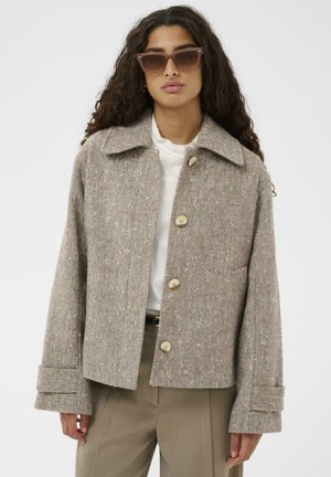 Braune Fischgräten-Blazer mit Kragen, fünf Knöpfen und breiten Manschetten, kombiniert mit einer weißen Bluse und olivgrünen Hosen.