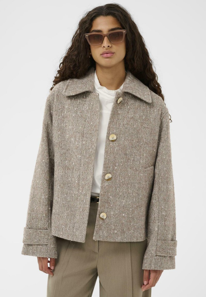 Rjav blazer s ribjo kostjo, ovratnikom, petimi gumbi in širokimi manšetami, kombiniran z belo bluzo in olivnimi hlačami.