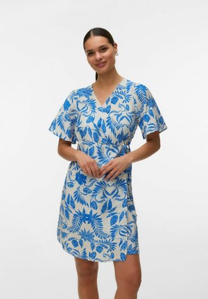 Kvinde med mørkt hår bundet tilbage iført en hvid knælængde kjole med blå blomsterprint og korte ærmer mod en ensartet hvid baggrund.